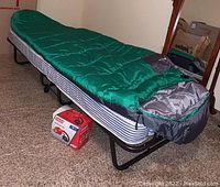 Folding twin bed frame with striped mattress pad, green sleeping bag on top, Coleman pump box underneath