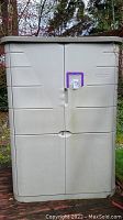 Front view of Rubbermaid plastic shed with double doors, beige color, showing slight dirt and wear from outdoor use on back deck.