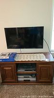 Front view of Samsung TV mounted on wooden TV stand with DVD players stored inside the shelves and storage compartments. A crocheted lace runner is placed on top of the stand with a box on it.