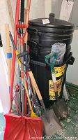 Photo showing various yard tools including shovels and rakes leaning against two large black plastic trash bins.