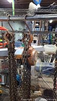 Front view of an orange chain hoist suspended from a metal rack with rusted chains and hooks attached, in a workshop environment.