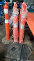 Seven orange delineator posts standing upright, some wrapped in plastic, with orange handles and two white reflective strips on each post. Four black rubber octagonal bases placed nearby.