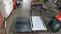 Top view of two dolly carts side by side on concrete floor, one with handle and silver platform, the other black flat platform with no handle.
