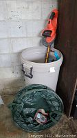 Photo showing the cordless Black & Decker Hedge Hog hedge trimmer resting inside a large Rubbermaid bin along with blue can grabbers, red and black digital water timer, and green collapsible leaf bag on the floor in front. The bin is approximately 31 inches tall.