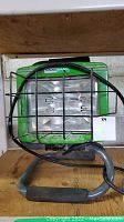Front view of green HaussMann portable work light showing protective metal grille, handle and power cord.