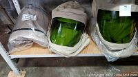 Three beige fabric-covered face shields with green arc flash visors placed on a shelf; one still in plastic packaging.