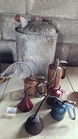Collection showing large cylindrical metal gas can with multiple smaller vintage metal oil cans around it, placed on floor against concrete block wall.
