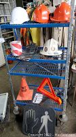 Metal shelf rack holding various safety gear including hard hats (white and orange), black gloves, caution light, traffic cone, safety triangle, and crosswalk sign.