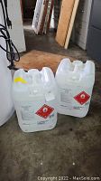 Two white 5-gallon plastic containers with hazardous material flammable liquid label UN1987 on side, placed on floor in an indoor setting.