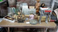 Table with various items including Coca-Cola tinted glassware, a wooden ship model, pasta maker, statues, votive candle holders, and other collectibles.