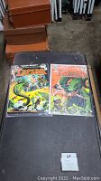 Two Tarzan comic books in plastic sleeves on black surface with brown suitcases in background