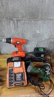Overview of cordless drills, battery chargers, and battery pack on wooden surface against concrete wall.