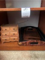 Photo of small wooden musical jewelry box with five drawers beside two wooden serving trays placed on shelf.