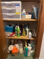 View of wooden closet shelves containing 3 drawer plastic storage cabinet, cleaning supplies, bottles, folded linens and other bathroom essentials.