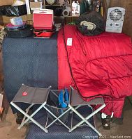 Photo shows red sleeping bag draped over couch, two gray folding camping chairs with metal frames in front, Playmate cooler in red with other camping gear stacked beside them.
