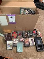 Photo of cassette tapes arranged in front of a cardboard box with some tapes inside the box and a portable cassette player on the right side.
