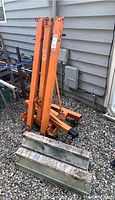 Metal engine hoist and portable sawhorse positioned side by side on gravel next to house siding. The engine hoist is folded down, orange color with visible lifting hook. The sawhorse has a wooden top marked by visible wear.