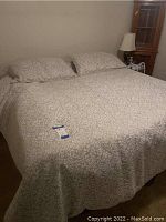 King-sized bed fully made with beige and white patterned bed linens and two pillows against a wall with a lamp and wooden corner cabinet visible