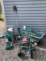 Front view of spoked wheeled garden hose reel with red hose, rear showing 'Professional' paint sign and other reels, tools and wheelbarrow in background