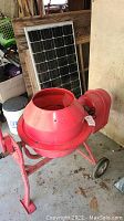 Red mini cement mixer with open drum and solar panel in background.