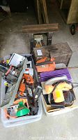 Overview of two plastic bins containing assorted tools, including orange chainsaw, hand saw, metal tool box, and other items.
