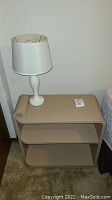 Beige night stand with two shelves and white lamp on top, next to bed.