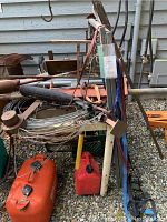 Photo of scrap metal including pipes, cables, hydraulic lift components, and fuel cans against a house exterior wall.