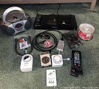 Overall view of all electronics and accessories included in lot, showing Sony DVD player, Lite-On DVD/CD-ROM attachment, remotes, cables, blank CD-R discs, portable stereo, and timers