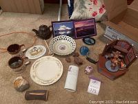 Photo shows an assortment of mugs, plates, postcards, rocks, porcelain decor, and wooden figurines arranged on carpet.
