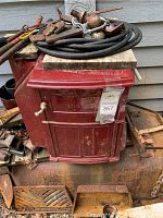 Encore red metal wood stove with parts piled on top and rusty metal pieces around base.