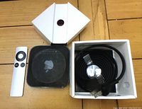 Photo showing Apple TV device, remote control, and cables in box on wood surface.