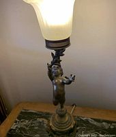 Full view of vintage bronze Art Nouveau / Deco table lamp with sculpted cherub figure and frosted glass shade illuminated.