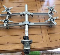 Full view of the quad monitor mount showing two horizontal arms with VESA mounting brackets, central vertical pole, and heavy-duty desk clamp base.