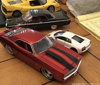 Photo shows four diecast cars, including a red muscle car with black stripes and white small scale sports car.