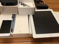 Lot contents showing Apple iPad, Apple TV units with remote, Apple iPod, boxes, and bag on wooden surface.