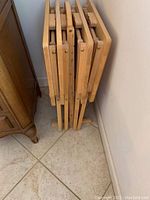Four light wood folding tables stacked vertically in folded position against a wall, showing the tables' thin profile and metal hinge details.