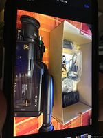 Sony video camera showing side with AF 60x zoom lens and branding. Next to box containing cables, remote control, and user manual in plastic bags.