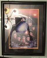 Framed Star Trek print called The First Family by Keith Birdsong in black frame with orange mat, glass front with reflection of ceiling light.
