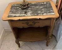 Front view of antique walnut nightstand with black and white marble top, single drawer with ornate handle, curved legs and lower shelf, placed on tiled floor.