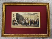 Framed print showing a courtyard scene with a crowd of people, mounted with a dark red matte and gold frame.