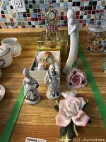 Photo showing enamelled decorative clock on gold stand, porcelain figurines (two women), porcelain woman holding child figurine, and porcelain flowers on table surface with mosaic tile background.