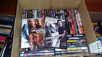 Box containing over 50 DVDs with visible action and adventure movie titles on top, some cases partially out of view within the box.