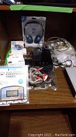 Photo of lot items displayed on wooden shelf including headphones in sealed packaging, cables and other electronics in plastic bags, various electronic cords bundled and loose.
