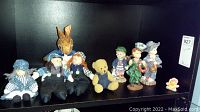 Collection of miniature dolls, teddy bear and children figurines on black shelf including cloth and ceramic/porcelain figures