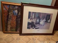 Two framed artworks leaning against wall on floor: one a ballet dancers print, other an autumn trees puzzle