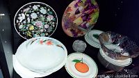 Overview photo showing stacked fruit plates with orchid and bird floral designs, oval painted bowl and several small plates, clear crystal coasters and colorful enamel plate