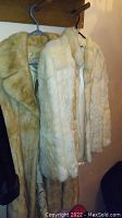 Two vintage coats hanging on wooden hooks, one medium beige fur coat and one light-colored rabbit fur coat.