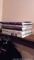 Stack of six hardcover books on a wooden shelf, titles visible for most books.