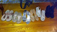 Seven pairs of ladies shoes arranged on wooden floor showing various styles including sandals, open-toe high heel, and boots.