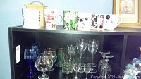 Shelf view showing a mix of blue wine glasses, clear snifter glasses, green imprinted glasses and six colorful printed mugs.
